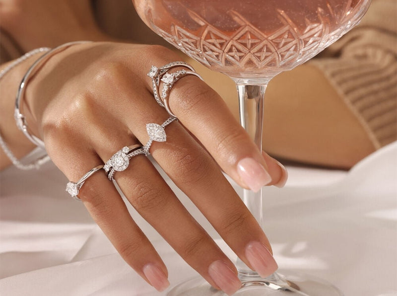 A woman with manicured nails wears multiple moissanite rings on her fingers, elegantly holding a crystal glass, showcasing the brilliance of the rings in a sophisticated setting.