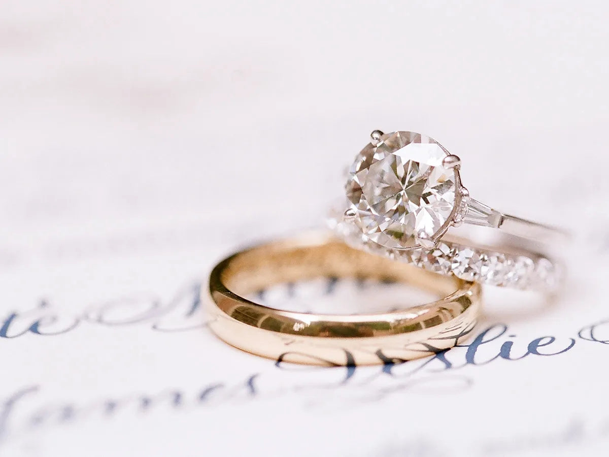 A gold wedding band and a diamond engagement ring rest on a handwritten wedding invitation.